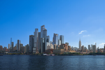 Obraz premium Modern skyscrapers of the Hudson Yards district and the Empire State Building reflecting in the Hudson River under a clear blue sky, Manhattan, New York City.