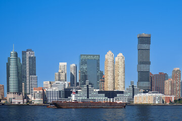 Obraz premium Panoramic view of the Jersey City skyline with modern glass skyscrapers reflecting sunlight under a clear blue sky, seen from the Hudson River.