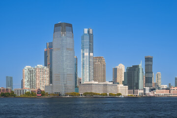 Obraz premium Panoramic view of the Jersey City skyline with modern glass skyscrapers reflecting sunlight under a clear blue sky, seen from the Hudson River.