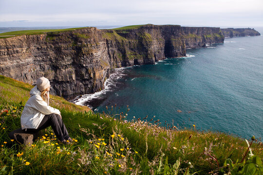 A solitary traveler sits amidst yellow wildflowers on a lush green cliffside, gazing out at the dramatic, rugged coastline of Cliffs of Moher and the vast blue Atlantic Ocean.