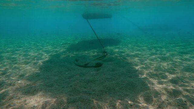 In the shadow of the boat, an anchor lies on a sandy bottom in shallow water, on a bright sunny day in the backlit sunlight