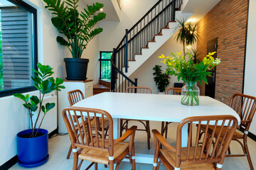 Fototapeta premium Modern dining room featuring empty table surrounded by six wooden chairs, large green potted plants placed around space, staircase and brick accent wall in background