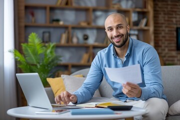 Young man smiling at camera while working on laptop, holding documents, calculating budget, and...