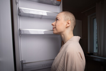 bald woman in a loose bathrobe looks thoughtfully at the inside of an open fridge in her apartment kitchen, symbol of food planning, healthy lifestyle and life after cancer treatment