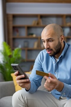 Man reacting with a shocked expression, holding a credit card and smartphone, experiencing financial stress from an unexpected banking error or online fraud while sitting at home