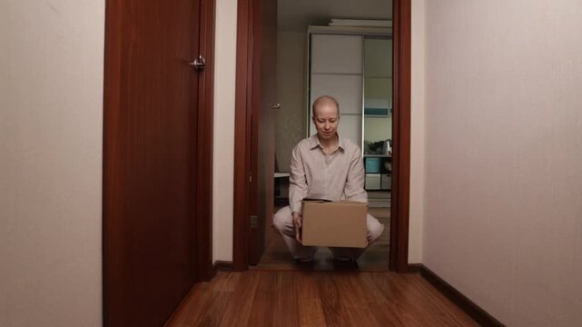 Oncology patient crouching on floor with cardboard parcel, home delivery of medicines and medical supplies, cancer support concept with woman receiving box at front door in modern flat.