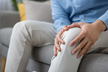 Middle-aged man sitting on a couch pressing his painful knee, grimacing from joint ache and inflammation, suggesting injury, arthritis or need for medical attention and treatment