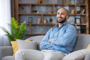 Relaxed smiling man with crossed arms on a cozy sofa at home, casually dressed and looking away,...