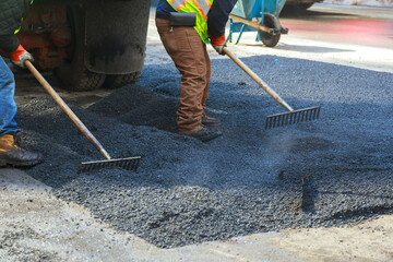 Two workers use rakes to spread asphalt on city street during road repair project