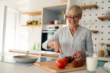 Fototapeta premium Smiling senior woman with short blonde hair cutting bread in bright modern kitchen at home
