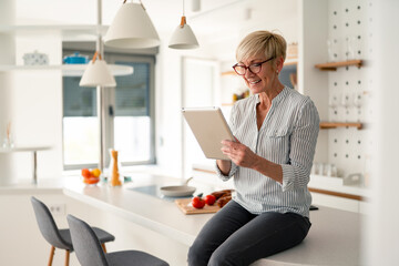 Happy senior woman wearing glasses working with tablet in bright modern kitchen enjoying morning work © Dorde