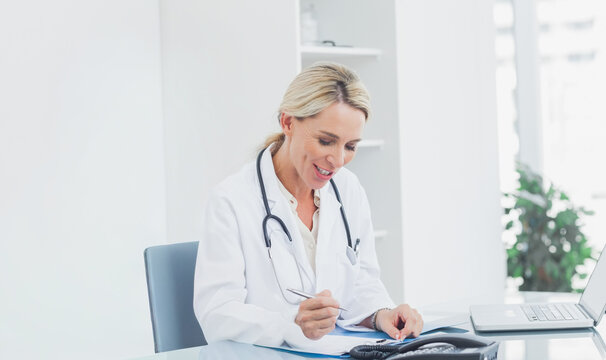 Mature adult female doctor sitting at clinic, writing files with pen and stethoscope on desk