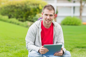 Man sitting on grassy lawn holding tablet, wearing red tee and gray hoodie, smiling at camera