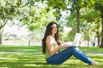 Adult woman sitting cross-legged on grassy lawn wearing sleeveless green top and jeans using laptop