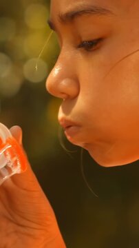 Vertical video: Lifting orange tub, child puffing and blowing to make bubbles in park, in peach top