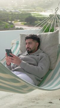 Vertical video: Opening eyes, man lying on balcony hammock checking texts on phone, grey sweater