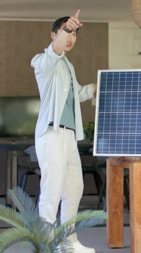 Vertical video: Raising arm, pointing, man in teal tee showing solar panel on wood stand at patio