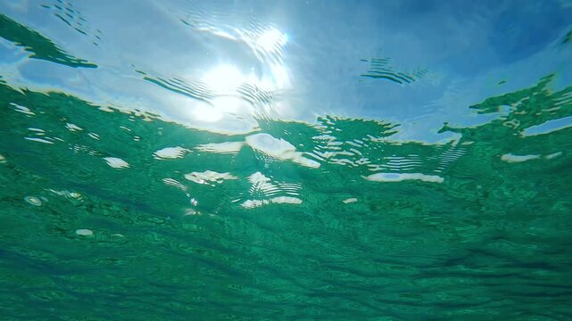 Weak breeze disperses ripples on the surface of emerald green crystal clear water in bright midday sun on the background of blue sky, backlit, underwater view