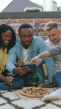 Adorable joyful 25s friendly diverse mates having fun together at revisioning interesting and amusing videos on phone during meeting in somebody's home on terrace,slow motion