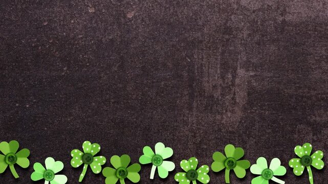 St Patricks Day bottom border of moving handmade paper button shamrocks. Stop motion loop video. Top view over a dark stone background with copy space.