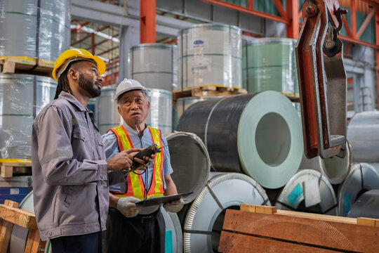 Two factory workers discussing metal coil inspection in an industrial warehouse. They wear safety gear, including helmets and vests, emphasizing teamwork, manufacturing, and industrial safety.