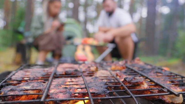 Delicious pork meat grilling on a barbecue rack over a campfire during a picnic in the woods, with a blurry couple of friends relaxing in the background and smoke rising from the hot charcoal