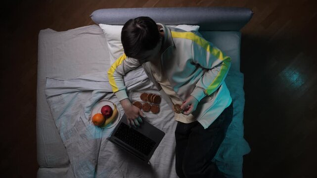 Young man lying in bed late at night, browsing a laptop and eating cookies, instead of focusing on productive work, representing procrastination and modern lifestyle