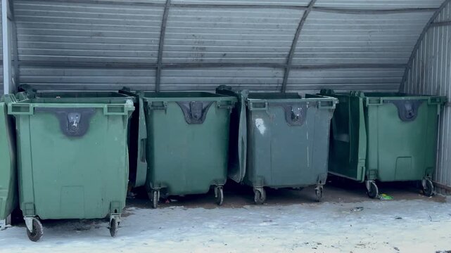 Dark green garbage bins in courtyard, lined under metal shelter on snow dusted concrete, empty wheels and closed lids, chipped paint and small stickers, quiet urban morning near apartment
