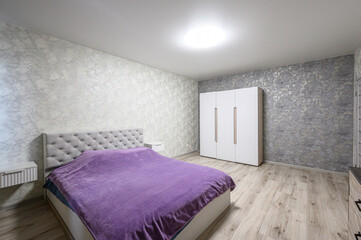 Fototapeta premium Spacious bedroom with double bed (purple blanket), tufted headboard, white wardrobe, light wood flooring, textured grey-white walls, minimalist decor, ceiling light.
