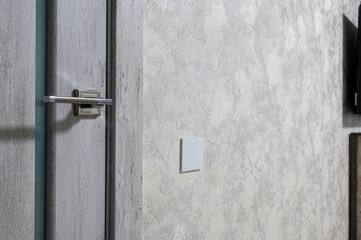 Light grey textured wall with modern square light switch, adjacent to light wood-finish door with...