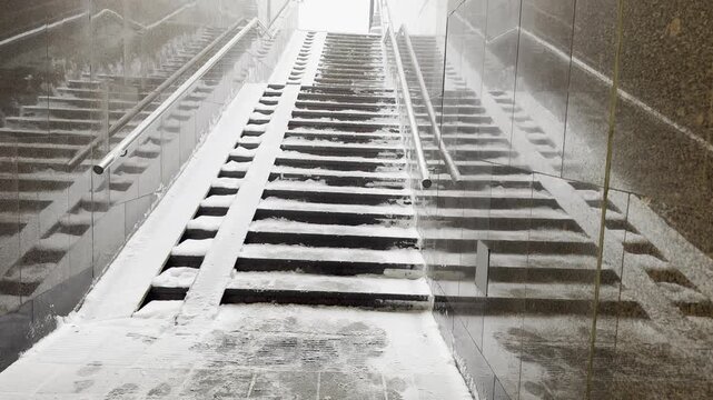 Symmetrical view of granite stairs and a disability ramp covered in fresh snow inside a city pedestrian underpass with polished dark stone walls.
