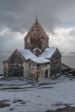 Stone church of Sevanavank Monastery rises above snowy shore of Lake Sevan in Armenia, dark volcanic masonry contrasts with a cold winter sky