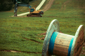 Large wooden cable spool wrapped in protective plastic resting on a grassy hillside. Preparations for infrastructure installation. On background, out of focus, a yellow excavator.
