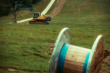In front out of focus large wooden cable spool resting on a grassy hillside. Preparations for infrastructure installation. a yellow excavator is working on background.
