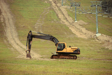 A large yellow hydraulic excavator parked on a grassy hillside. Undergoing construction or land modification. Preparing terrain for snowmaking infrastructure