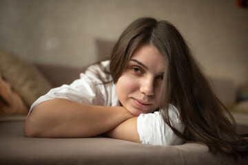 Fototapeta premium Young woman with long dark hair resting on a couch, wearing a white shirt, with soft pillows and a neutral background creating a cozy indoor atmosphere