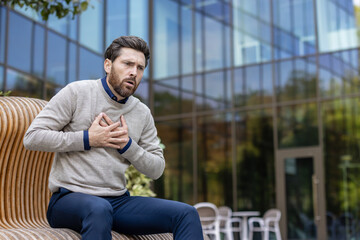 A young businessman sits on a bench near an office center and holds his hands to his chest, feeling...