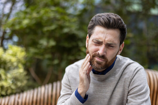 Close-up photo of a young man sitting on a bench, holding his cheek with his hand and suffering from a toothache