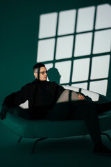 Fototapeta premium A portrait photo of a male model in a dark studio with window light. The handsome man wearing glasses looks into the camera. The businessman and his fashionable, elegant style