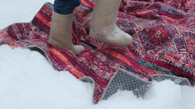 Close-up of a woman beating and cleaning a rug with a broom outdoors on fresh snow. Snow flies into the air during winter housekeeping and traditional carpet cleaning, with focus on boots, legs and ru
