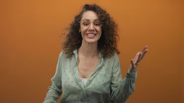 Woman wearing green polka dot blouse and glasses smiling with raised hand gesture in studio with orange backdrop; excitement playfulness warmth.