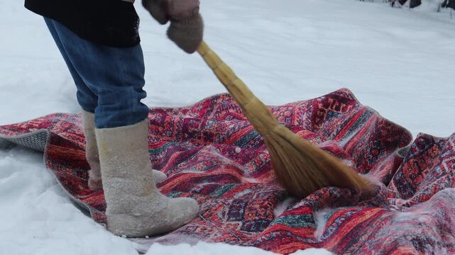 Close-up of a woman beating and cleaning a rug with a broom outdoors on fresh snow. Snow flies into the air during winter housekeeping and traditional carpet cleaning, with focus on boots, legs and ru