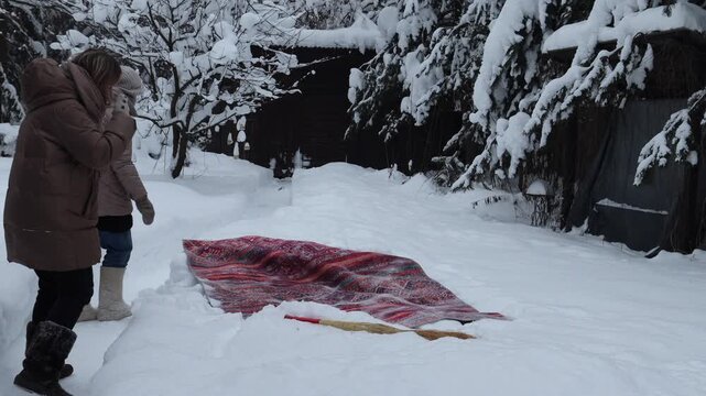 Close-up footage of a woman cleaning and beating a rug with a broom outdoors on fresh snow. Winter housekeeping and traditional carpet cleaning, with snow flying in the air and focus on boots, legs an