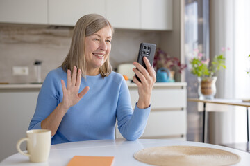 Senior woman at home smiling and waving during a smartphone video call, enjoying a virtual chat...