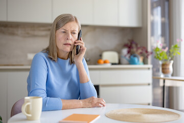 Elderly woman in kitchen listens intently on her mobile phone, worried and pensive during a...