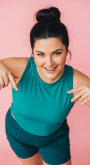 Smiling woman in teal sportswear against pink background, pointing downwards, showcasing product placement opportunity