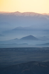 Aerial view from high spot of valley at front surrounded by hills covered in fog, church located on top of hill and main Causasus mountains in Georgia at sunset on background, idyllic winter landscape