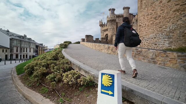 Female pilgrim carrying a backpack while walking the Way of St. James, passing a scallop shell signpost with the historic Templar Castle of Ponferrada looming in the background