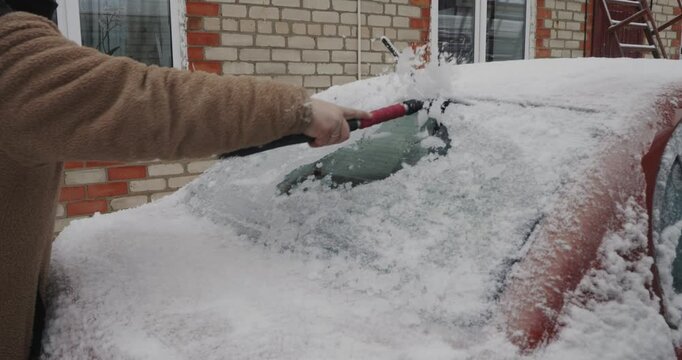 Person in a warm jacket cleaning a thick layer of fresh snow from the windshield of a red car using a special brush and scraper tool after a heavy snowfall on a cold winter day.