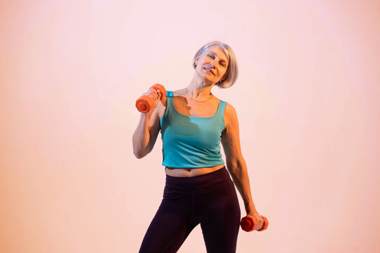 Confident mature woman training with dumbbells in a modern gym. Athletic middle-aged female in sportswear holding weights and preparing for strength training. Concept of active aging, healthy lifestyl